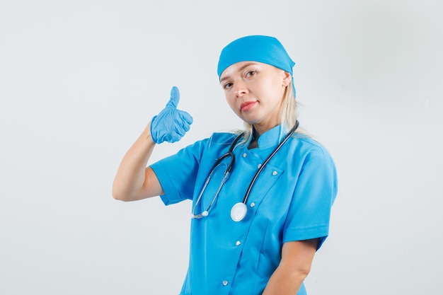 파란색 유니폼을 입고 엄지 손가락을 보여주는 여성 의사가 기쁘게 생각합니다.