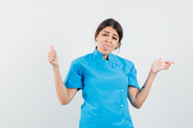 Female doctor showing thumb up, pointing aside, sticking out tongue in blue uniform