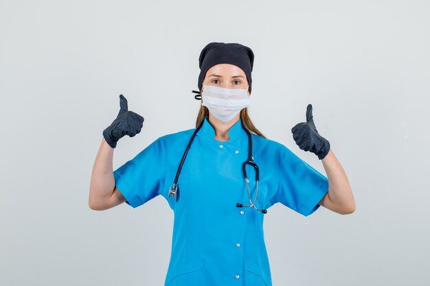 Female doctor showing thumbs up in uniform, gloves, mask and looking pleased. front view.