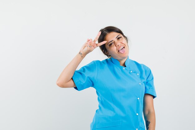 Female doctor showing v-sign, sticking out tongue in blue uniform and looking crazy