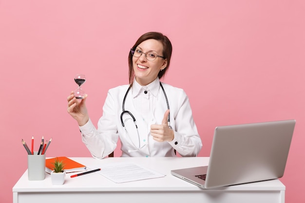 여성 의사는 파스텔 핑크색 벽 배경에 격리된 병원에서 의료 문서를 들고 있는 컴퓨터 책상에 앉아 있습니다. 의료 가운 안경 청진 기에서 여자입니다. 의료 의학 개념입니다.
