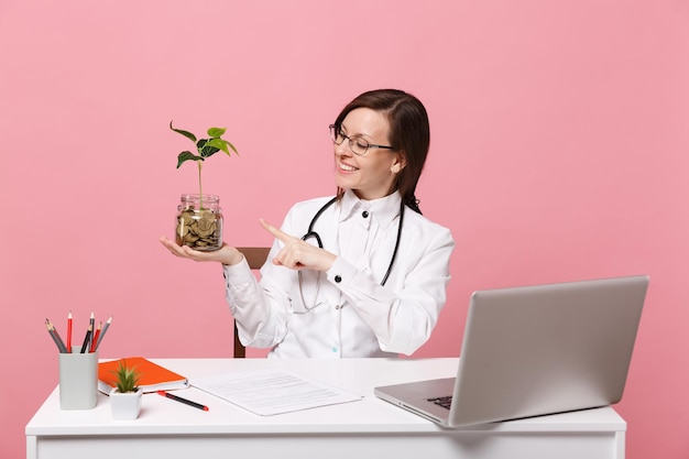 여성 의사는 파스텔 핑크색 벽 배경에 격리된 병원에서 의료 문서와 동전을 들고 컴퓨터 책상에 앉아 있습니다. 의료 가운 안경 청진 기에서 여자입니다. 의료 의학 개념입니다. 프리미엄 사진
