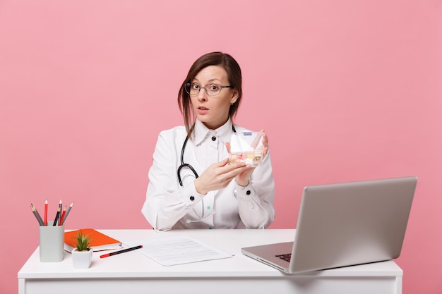 Женщина-врач сидит за столом и работает на компьютере с таблетками в больнице, изолированными на пастельных розовых стенах. женщина в медицинском стетоскопе очков мантии. концепция медицины здравоохранения.