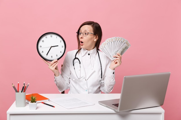 Female doctor sit at desk work on computer with medical document hold money in hospital isolated on pastel pink wall background. woman in medical gown glasses stethoscope. healthcare medicine concept.
