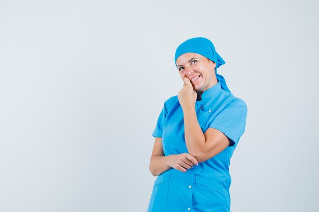 Женщина-врач, стоя в позе мышления в синей форме и выглядела счастливой, вид спереди.