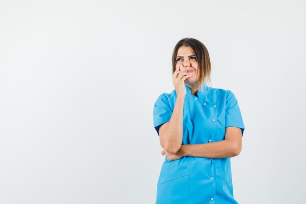 Женщина-врач, стоящая в позе мышления в синей форме и нерешительно выглядящая