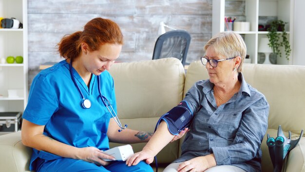 Female doctor taking blood pressure of senior woman in nursing home sitting on sofa