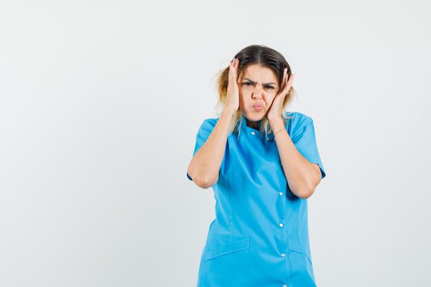 Female doctor touching face skin, pouting lips in blue uniform and looking gloomy
