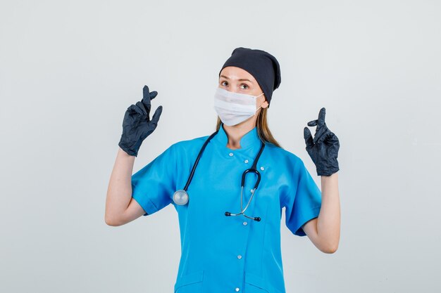 Female doctor in uniform, gloves, mask crossing fingers and looking hopeful