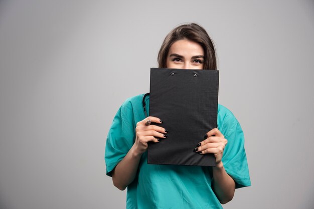 Female doctor in uniform hiding behind clipboard. 