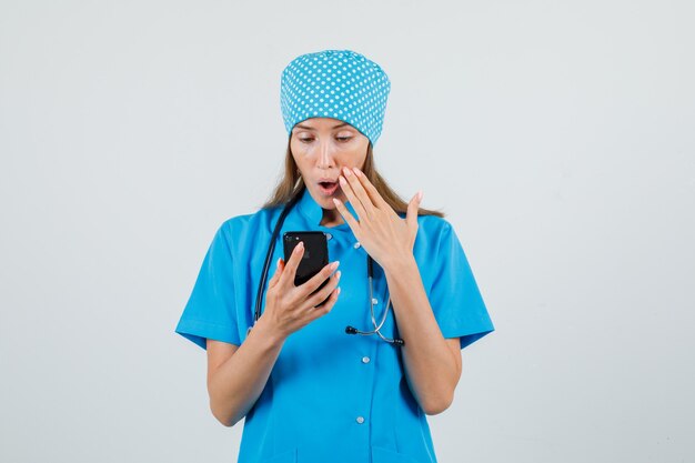 Medico femminile che utilizza smartphone in uniforme blu e che sembra sorpreso