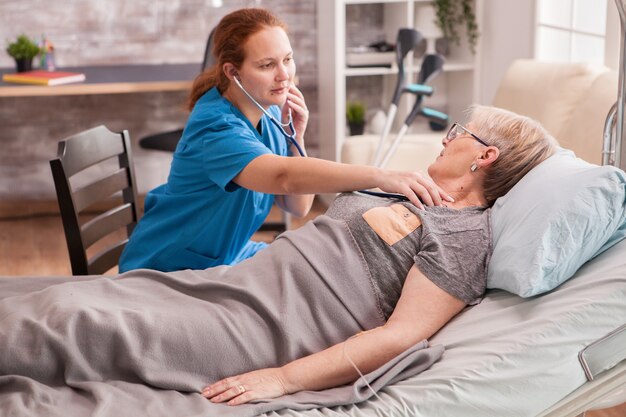 Female doctor using stethoscope to check old woman heart in nursing home.