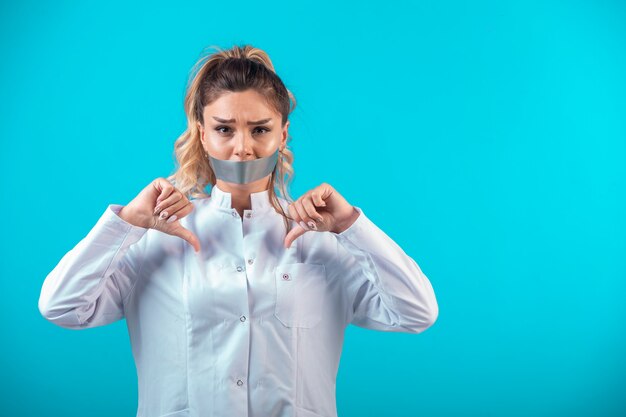 Female doctor in white uniform covering her mouth and makes thumbs down.