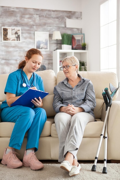 Female doctor with senior woman sitting on couch in nursing home writing a prescription on clipboard.