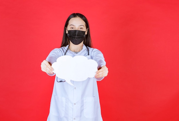 Female doctor with stethoscope and in black mask holding a cloud shape blank info desk. 