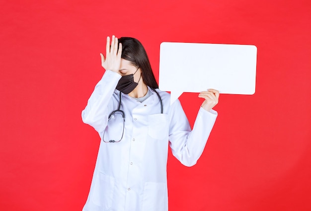 Женщина-врач со стетоскопом и в черной маске держит прямоугольный информационный стол и держит голову
