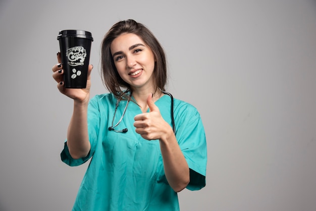 Женщина-врач со стетоскопом держит кофе и дает большие пальцы руки.