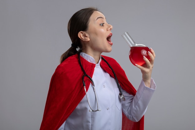 흰색 의료 유니폼과 빨간 슈퍼 히어로 케이프에 청진 기 여성 의사 붉은 액체와 함께 플라스크를 들고 흰 벽 위에 서서 마실 것입니다