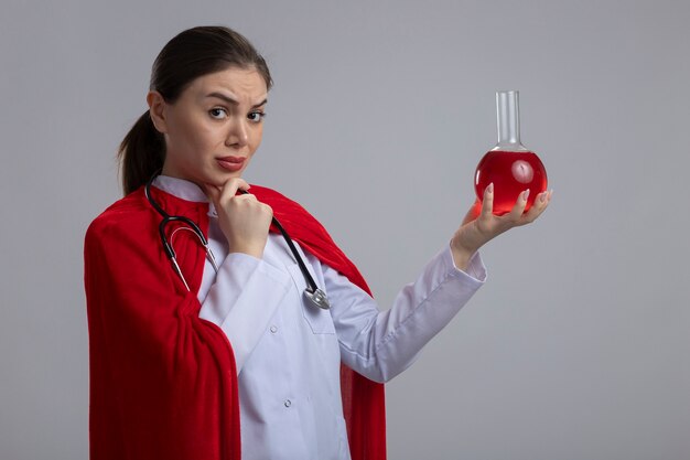 Женщина-врач со стетоскопом в белой медицинской форме и красной накидке супергероя, держащая фляжку с красной жидкостью, смотрит вперед с задумчивым выражением лица, думая, стоя над белой стеной