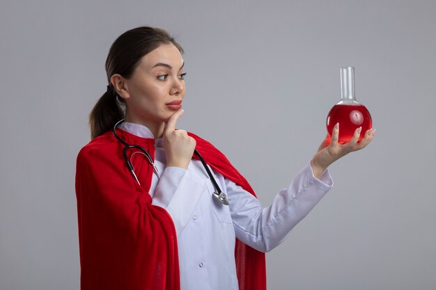 Female doctor with stethoscope in white medical uniform and red superhero cape holding flask with red liquid looking at it with serious face thinking standing over white wall