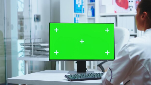 개인 클리닉 캐비닛에 녹색 화면이 있는 컴퓨터 작업을 하는 여성 의사. 유니폼을 입은 어시스턴트. 환자 진단을 확인하기 위해 클리닉 캐비닛에 크로마 키가 있는 모니터에서 작업하는 흰색 코트의 메딕.