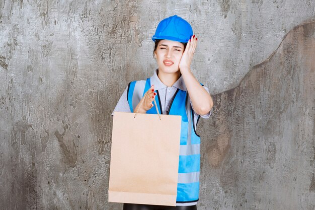 Female engineer in blue helmet and gear holding a cardboard shopping bag, holding head and looks tired