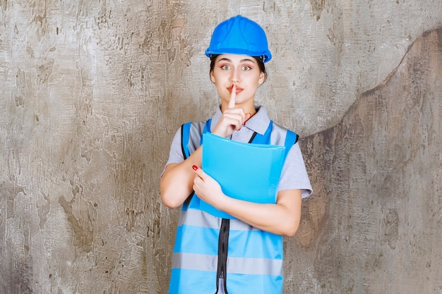 Female engineer in blue uniform and helmet holding a blue report folder and asking for silence.