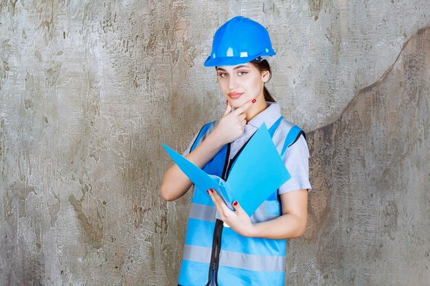 Female engineer in blue uniform and helmet holding a blue report folder and looks thoughtful.