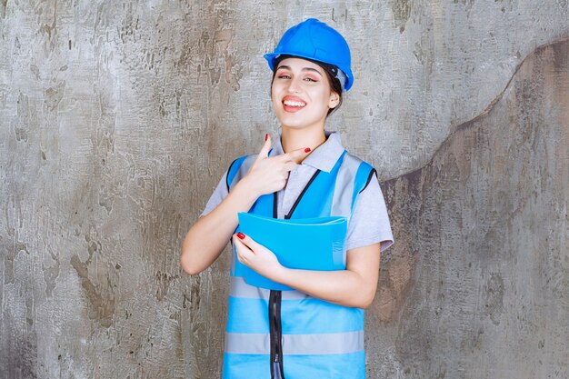 Female engineer in blue uniform and helmet holding a blue report folder and pointing at someone around