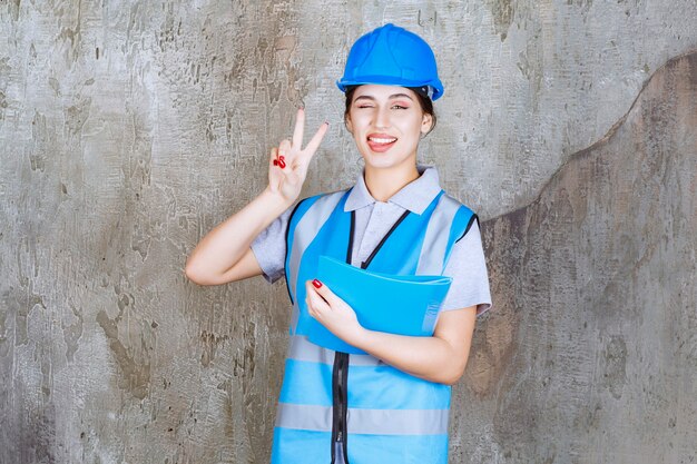 Female engineer in blue uniform and helmet holding a blue report folder and showing positive hand sign.