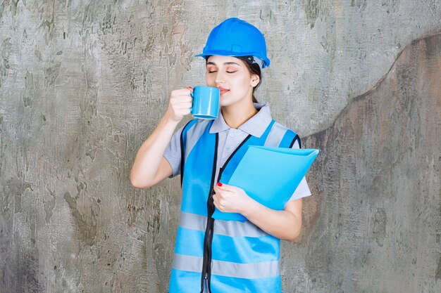 파란색 유니폼과 헬멧을 쓴 여성 엔지니어는 파란색 찻잔과 파란색 보고서 폴더를 들고 제품 냄새를 맡고 있습니다.