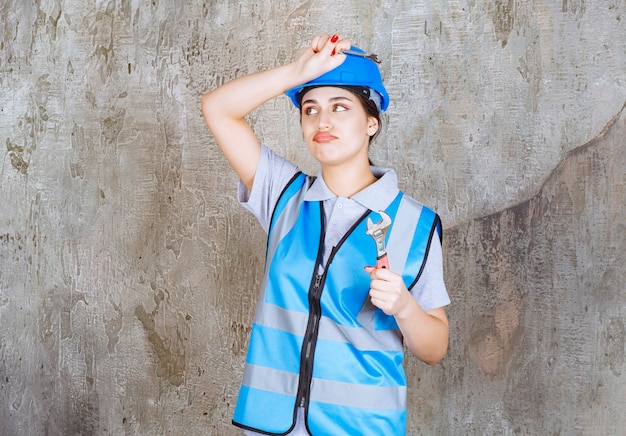 Female engineer in blue uniform and helmet holding a metallic wrench and feeling confused and thoughtful of how to use it.