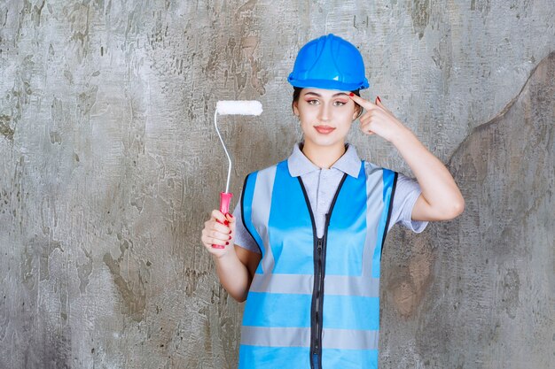 Female engineer in blue uniform and helmet holding a trim roller for painting and thinking and planning.