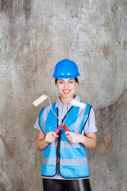 파란색 유니폼을 입은 여성 엔지니어와 페인팅을 위한 트림 롤러를 들고 있는 헬멧.