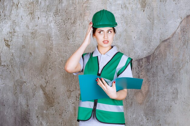파란색 폴더를 들고 녹색 헬멧을 쓴 여성 엔지니어는 혼란스럽고 사려깊게 보입니다.