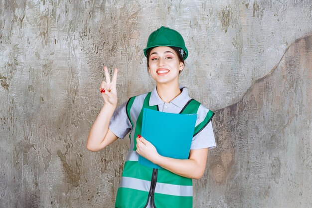 Ingegnere femminile in casco verde che tiene una cartella blu e che mostra il segno positivo della mano