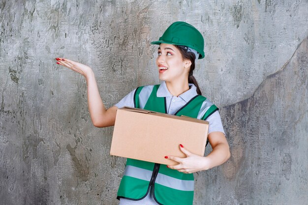 Female engineer in green helmet holding a cardboard box and pointing at someone around