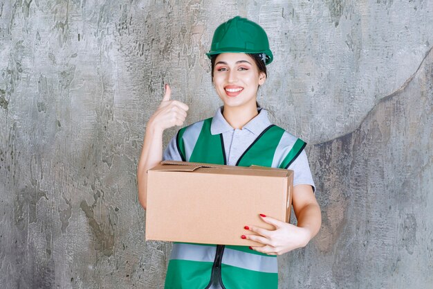 Female engineer in green helmet holding a cardboard box and showing satisfaction sign