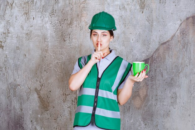녹색 헬멧을 쓴 여성 엔지니어가 녹색 커피잔을 들고 침묵을 요구합니다.