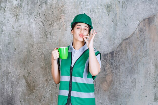 Женщина-инженер в зеленом шлеме держит кружку зеленого кофе и показывает знак удовольствия