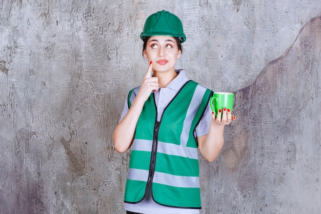 Foto gratuita ingegnere femminile in casco verde che tiene una tazza da caffè verde e che pensa