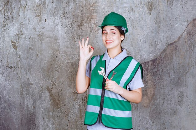 Female engineer in green helmet holding metallic wrench for a repair work and showing positive hand sign