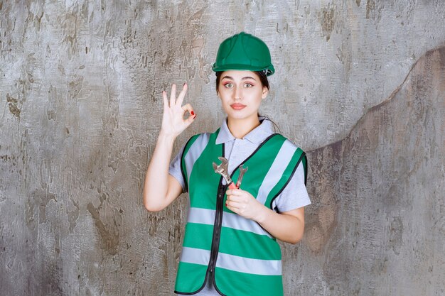 Female engineer in green helmet holding metallic wrench for a repair work and showing positive hand sign. 