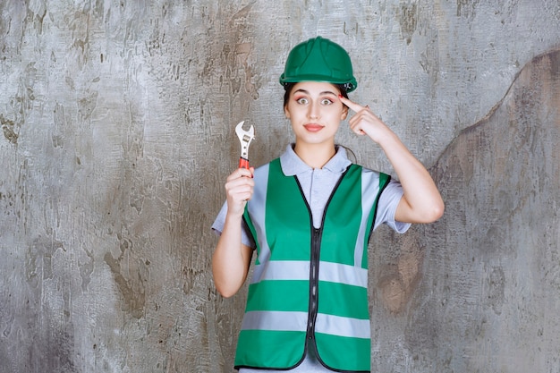 Female engineer in green helmet holding metallic wrench for a repair work and thinking about new ideas