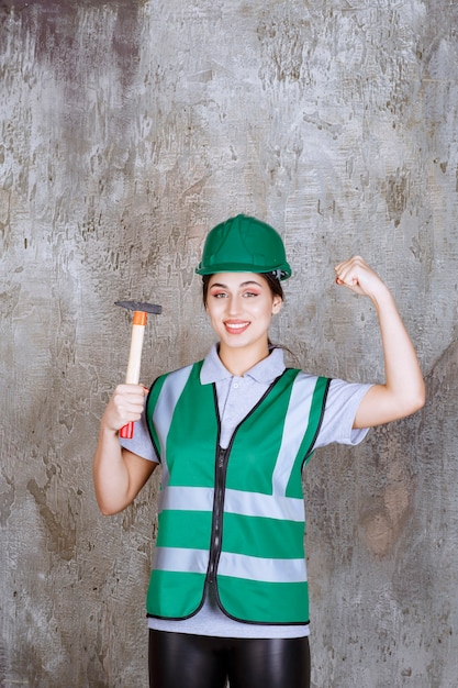 Female engineer in green helmet holding a wooden handled ax for a repair work and showing her arm muscles.