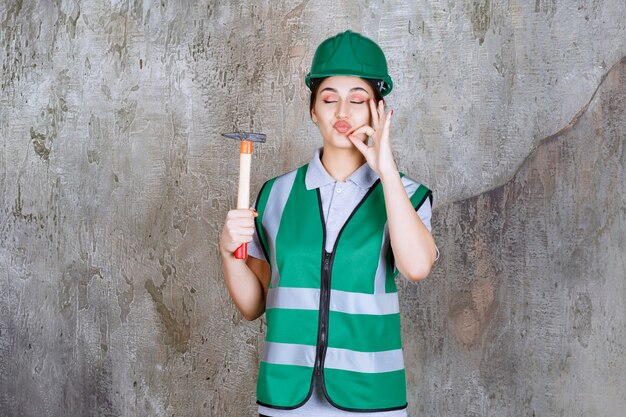 Женщина-инженер в зеленом шлеме держит топор с деревянной ручкой для ремонтных работ и показывает положительный знак рукой.