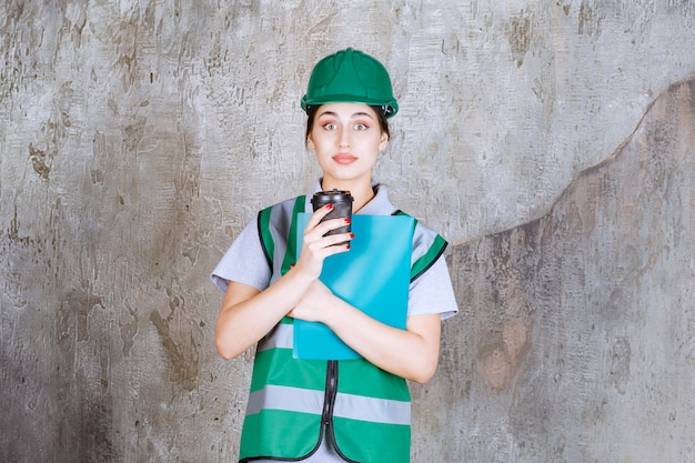 Ingegnere femminile in uniforme verde e casco che tiene una tazza di caffè nero e una cartella del progetto blu.