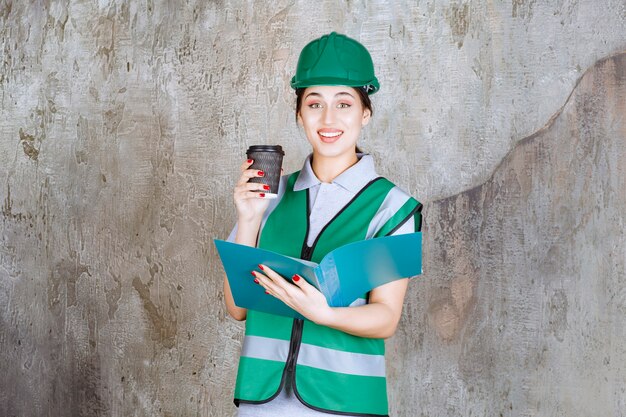 黒のコーヒーカップと青のプロジェクトフォルダーを保持している緑の制服とヘルメットの女性エンジニア。