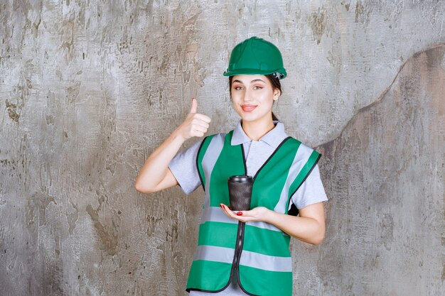 緑の制服と黒いコーヒーカップを保持し、製品を楽しんでいるヘルメットの女性エンジニア