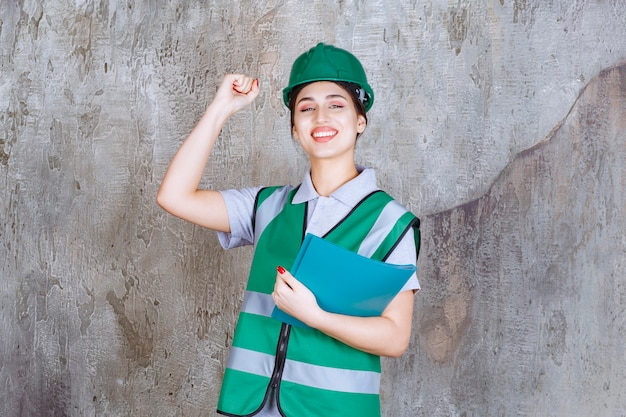 Ingegnere femminile in uniforme verde e casco che tiene una cartella di progetto blu e che mostra il segno positivo della mano.